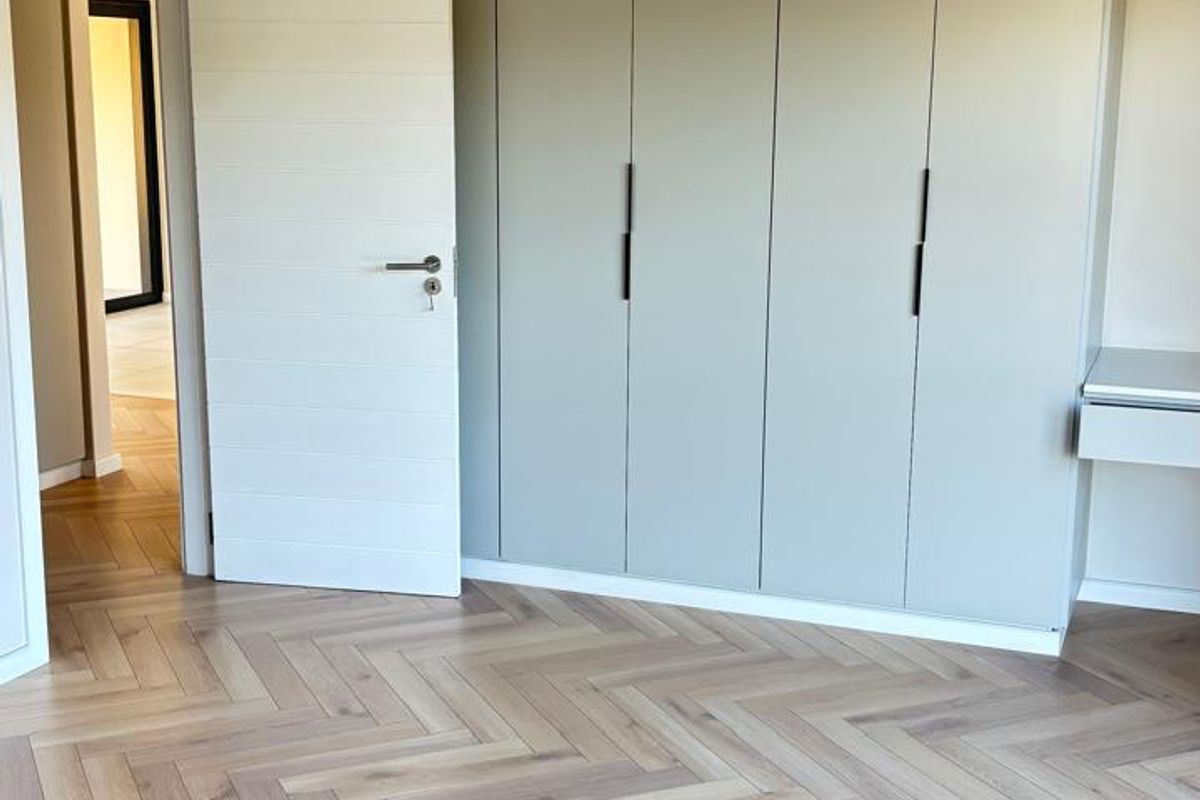 A bright, modern bedroom featuring high-quality herringbone timber flooring and extensive built-in cabinetry with a neutral finish.
