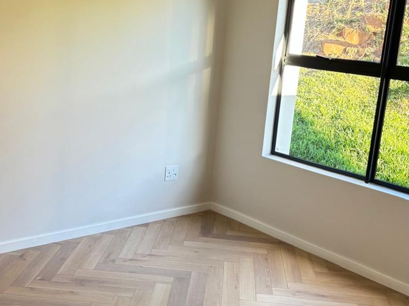 A bright, empty bedroom featuring modern herringbone timber flooring, neutral walls, and a large window with a black aluminium frame overlooking the garden.