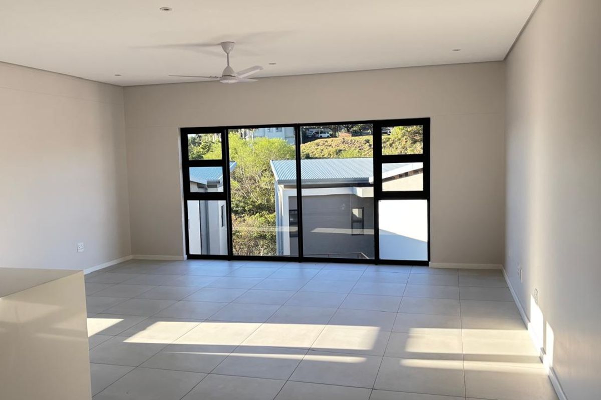 A bright, spacious, and empty living area featuring modern tiled flooring, a ceiling fan, and large windows with a view of the surrounding neighborhood.