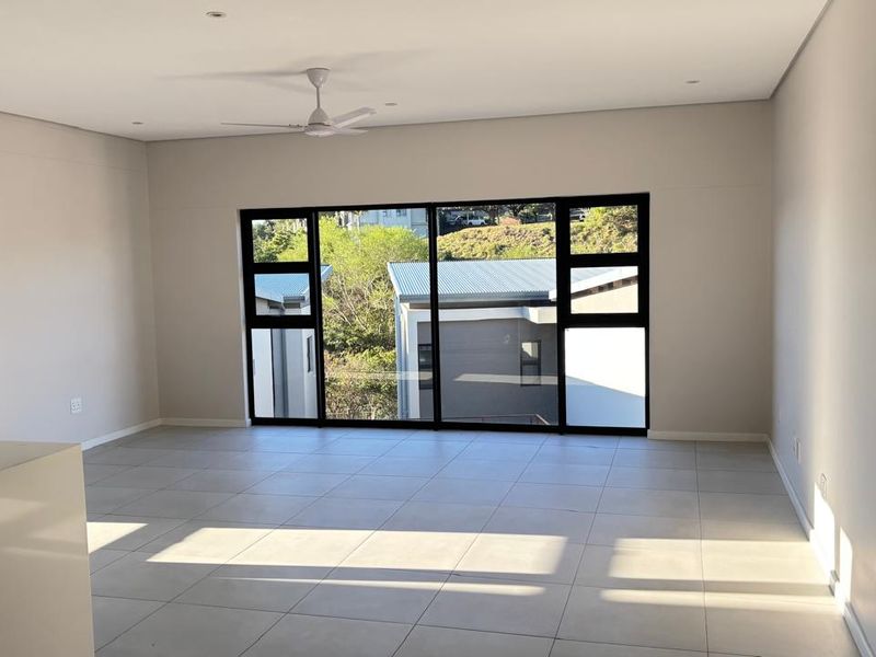 A bright, spacious, and empty living area featuring modern tiled flooring, a ceiling fan, and large windows with a view of the surrounding neighborhood.