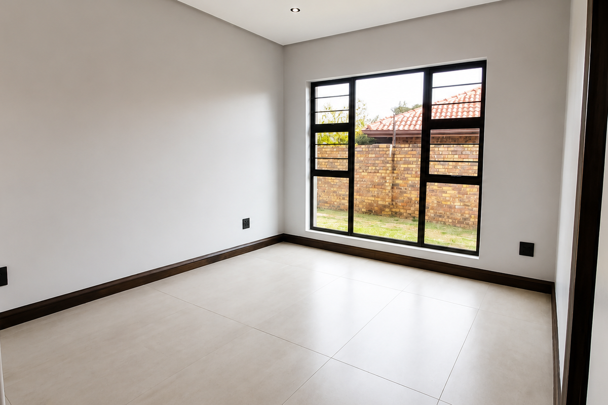 A bright, empty bedroom featuring neutral tiled flooring, modern black aluminium window frames, and recessed downlighting, offering a clean and versatile living space.