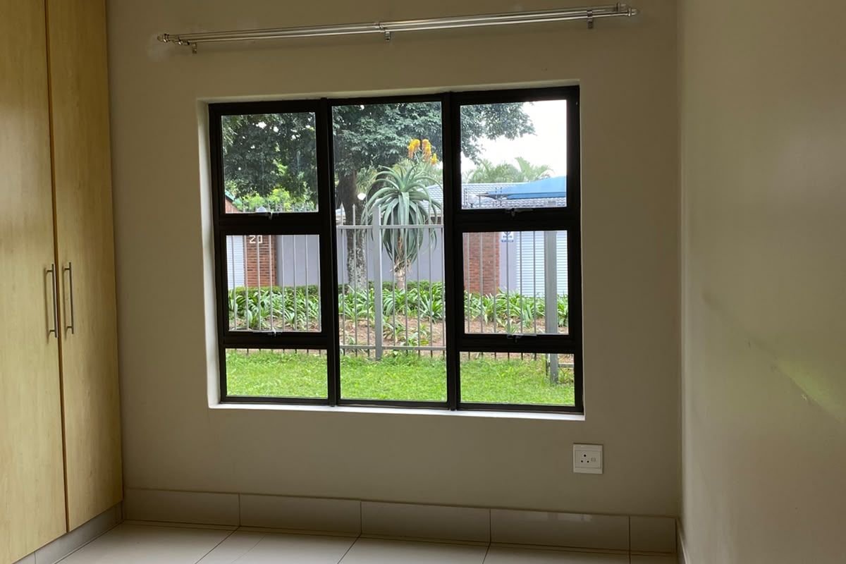 A bright, empty bedroom featuring neutral tiled flooring, built-in wooden cupboards, and a large window overlooking the garden area.