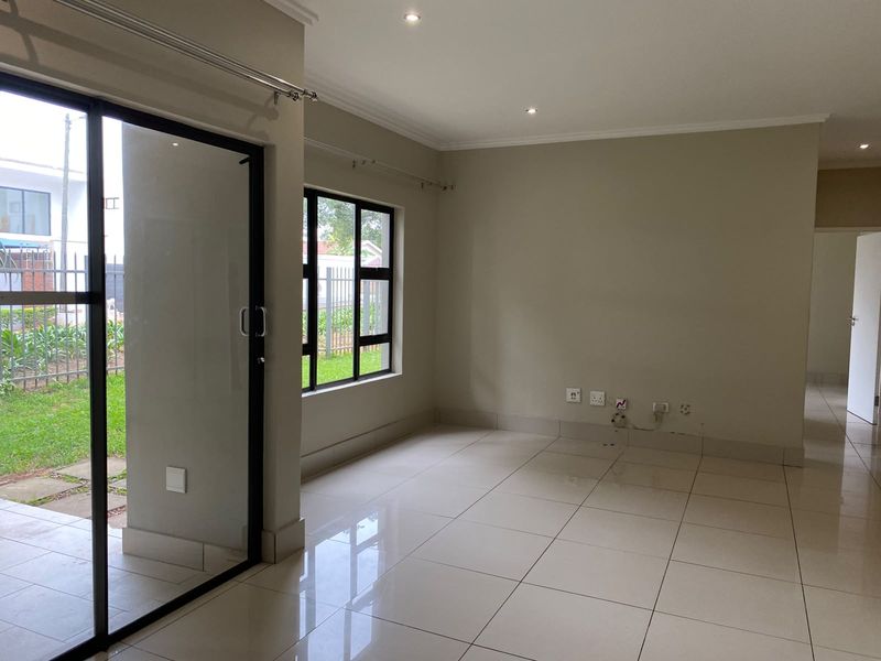 A bright, empty living room featuring neutral tiled flooring, recessed downlights, and large sliding glass doors providing access to the garden area.