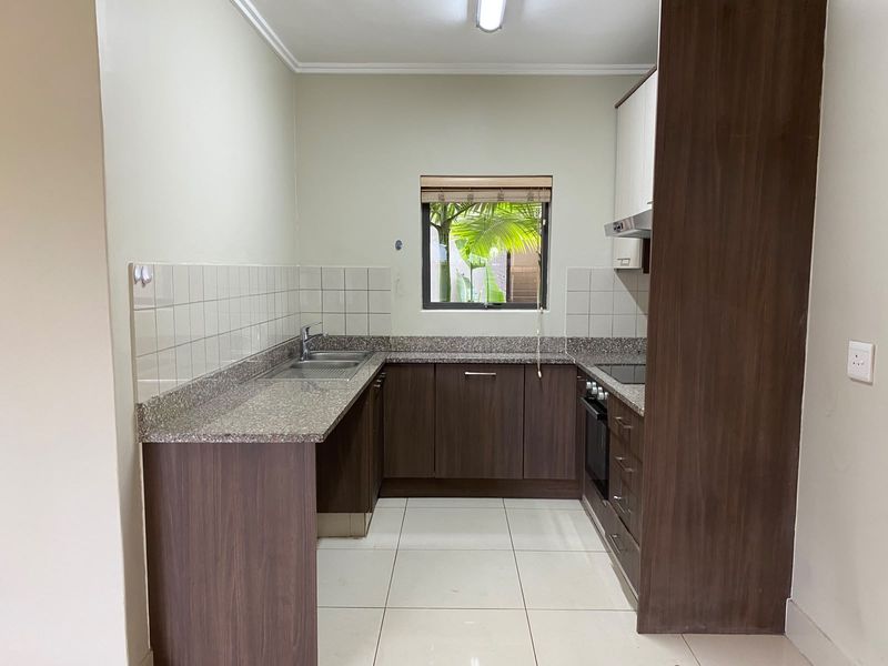 A clean, functional kitchen featuring dark wood cabinetry, granite countertops, a built-in oven, hob, and extractor fan with neutral tiled flooring.