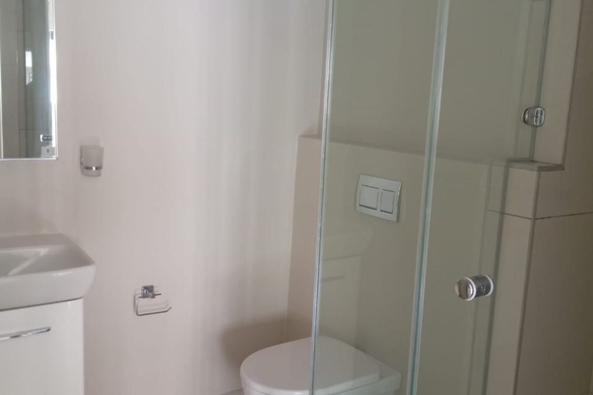 A clean, modern bathroom featuring a wall-mounted toilet, a glass-enclosed shower, and a vanity basin with neutral floor and wall tiling.