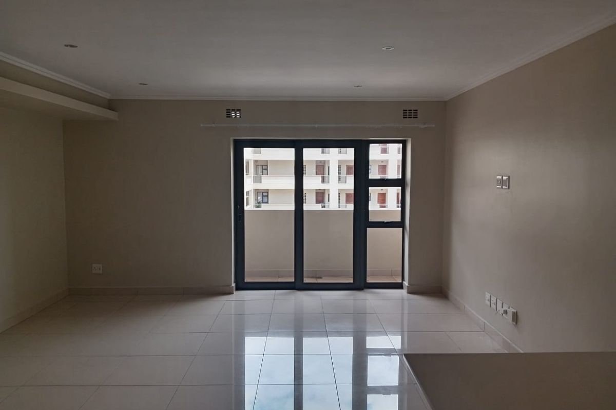 A spacious, empty living area featuring neutral tiled flooring, recessed lighting, and a large sliding glass door providing access to an outdoor balcony.