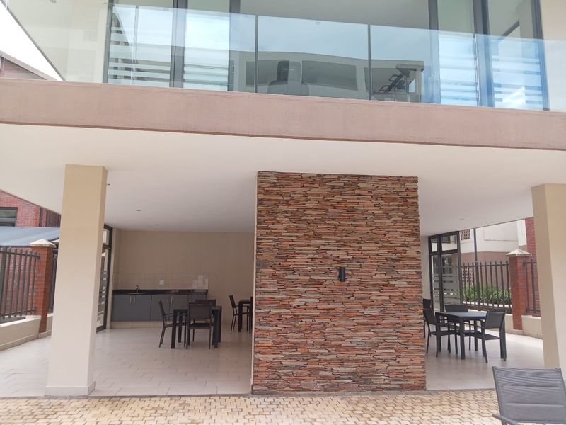 Covered outdoor patio area featuring a stone-clad feature wall, built-in kitchenette with sink, and tiled flooring, overlooking a modern apartment balcony.