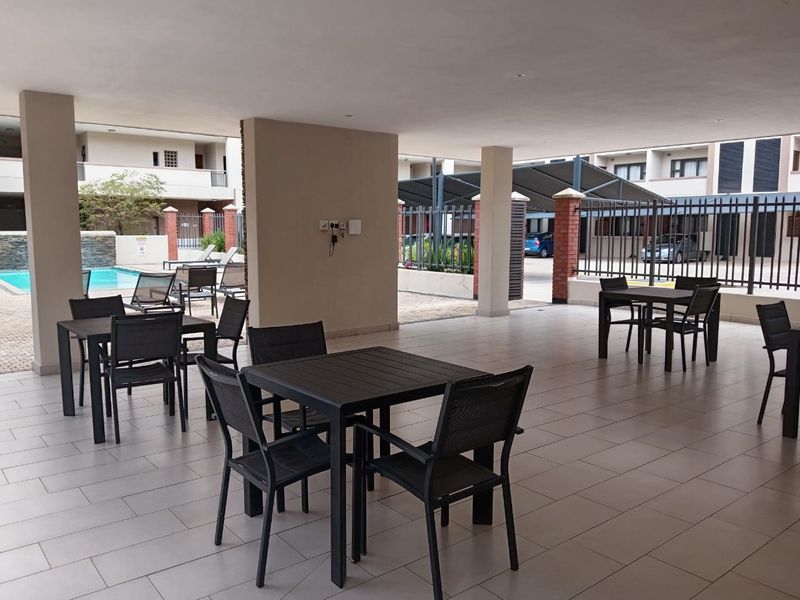 A spacious covered outdoor patio area featuring tiled flooring, brick pillars, and direct access to the complex pool and parking facilities.