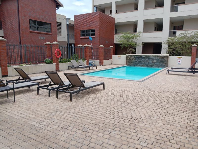 Communal swimming pool area featuring a stone water feature, paved deck, and brick building surrounds within an apartment complex.