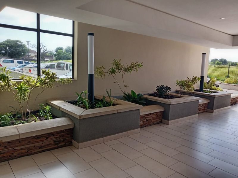 A covered outdoor patio area featuring built-in stone-clad planters, tiled flooring, and modern bollard lighting, overlooking a parking area.