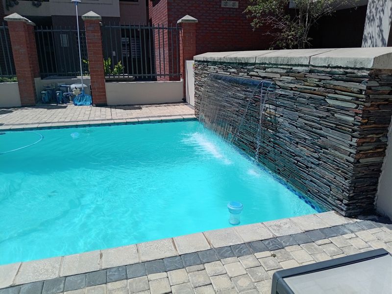 A well-maintained swimming pool area featuring a decorative stone water feature and paved surround within a secure complex.