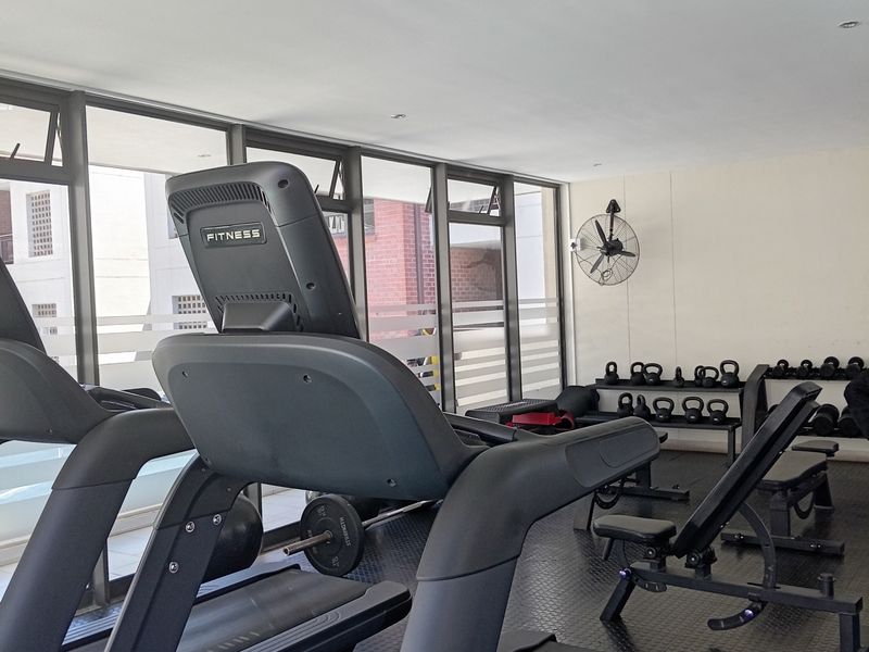 A well-maintained apartment gym facility featuring rubber flooring, large windows for natural light, and a wall-mounted industrial fan.