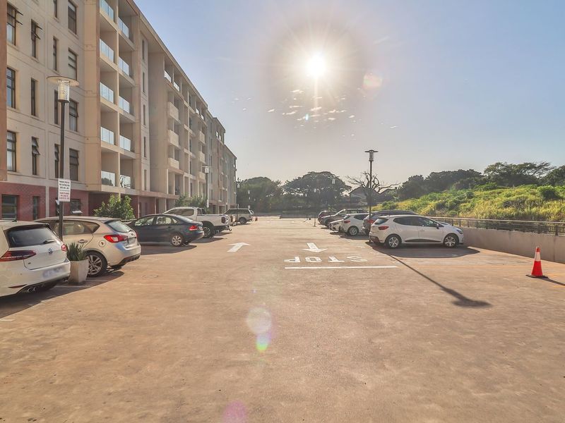 An outdoor parking area adjacent to a modern apartment building with glass balcony railings and paved surfaces.