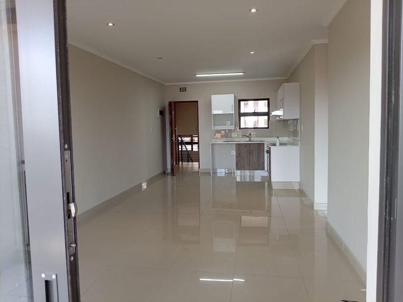An open-plan living and kitchen area featuring neutral tiled flooring, modern recessed lighting, and clean, contemporary kitchen cabinetry with a stainless steel sink.