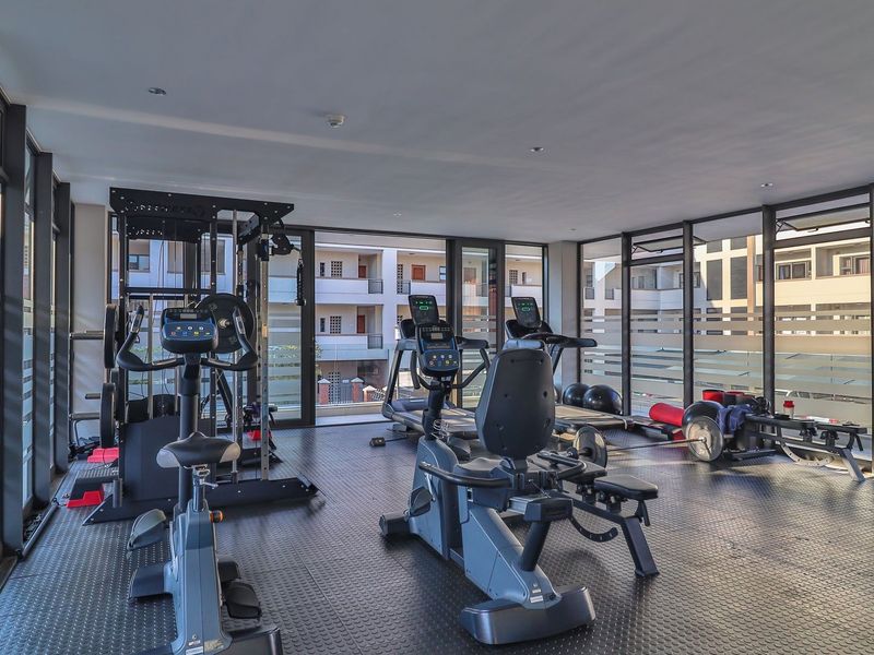 A well-lit apartment gym featuring durable rubberized flooring and expansive floor-to-ceiling windows providing natural light and views of the surrounding complex.