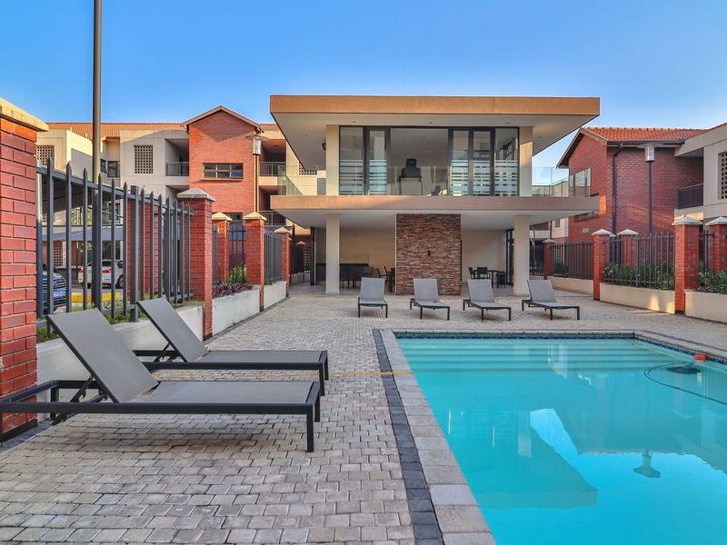 A well-maintained communal pool area featuring a paved deck, stone feature wall, and covered patio space within a brick-walled apartment complex.