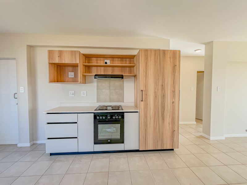 A modern, compact kitchen featuring light wood-tone cabinetry, a built-in oven with electric hob, extractor fan, and neutral tiled flooring.