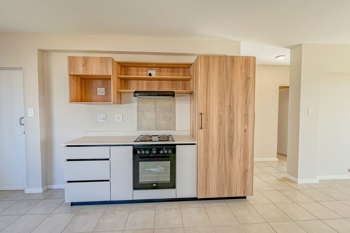 A modern, compact kitchen featuring light wood-tone cabinetry, a built-in oven with electric hob, extractor fan, and neutral tiled flooring.