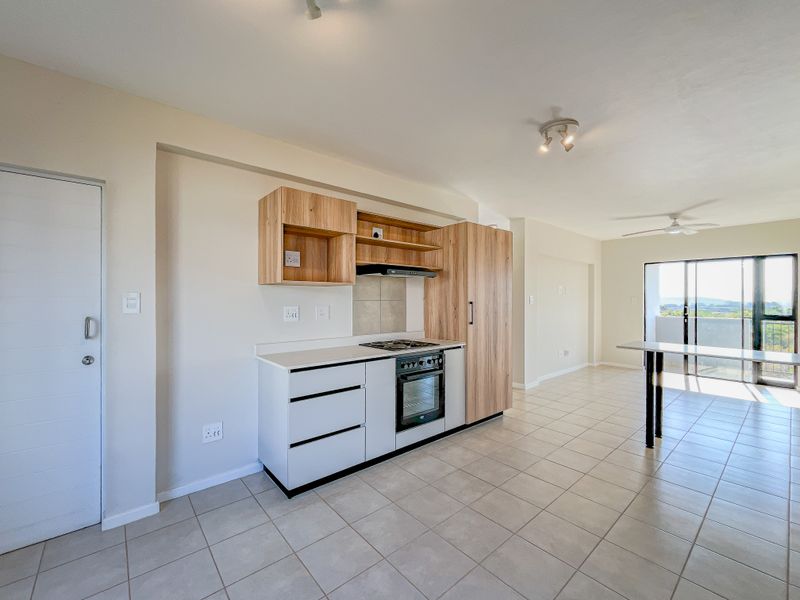 Modern open-plan kitchen and living area featuring light wood cabinetry, stone countertops, built-in appliances, tiled flooring, and access to a balcony.