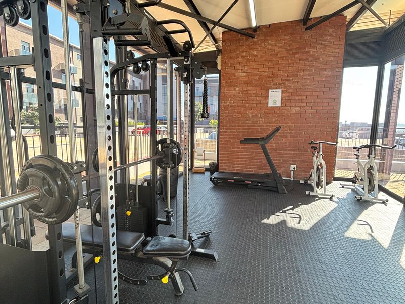 A modern, well-lit gym facility featuring exposed brickwork, industrial steel roof beams, durable rubber flooring, and expansive glass windows overlooking the complex.