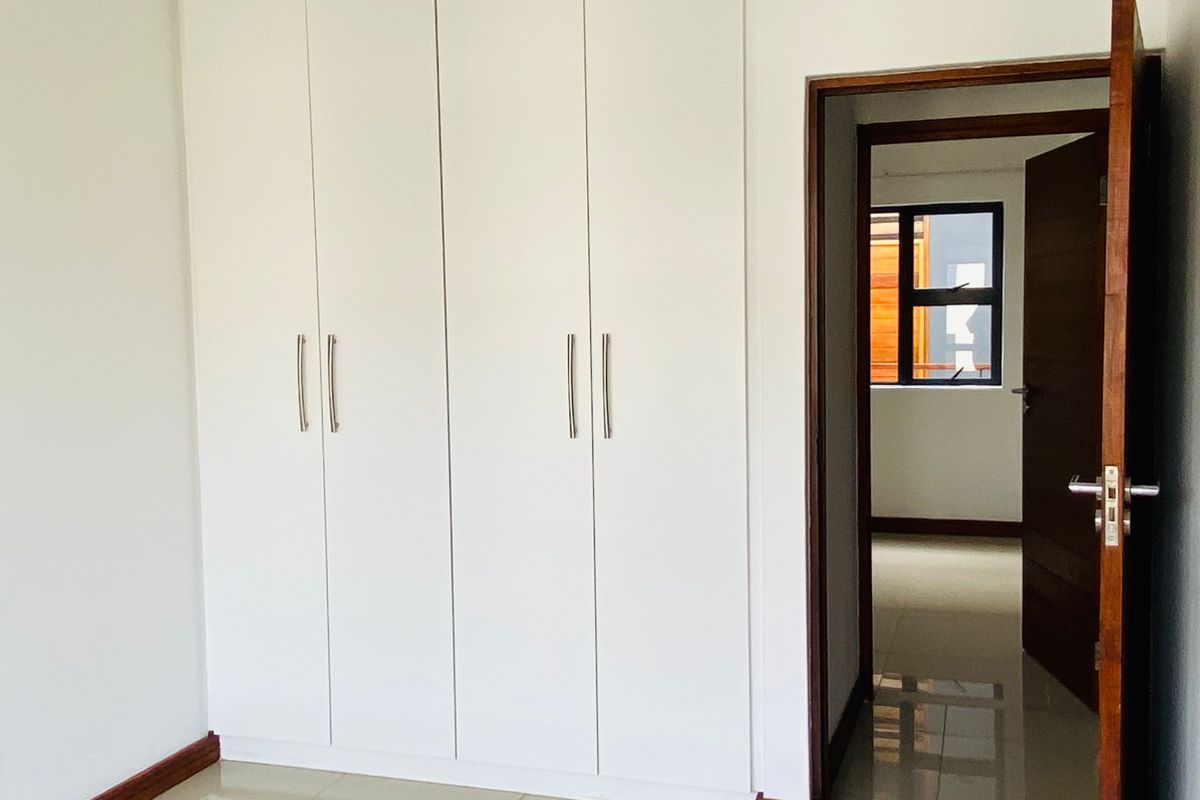 A clean, empty bedroom featuring modern white built-in cupboards, neutral tiled flooring, and a wooden door frame leading to an adjacent room.