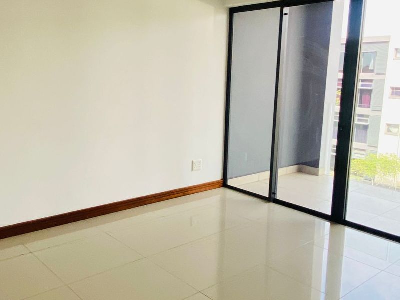 A bright, empty living area featuring neutral tiled flooring and a large sliding glass door providing access to an outdoor balcony.