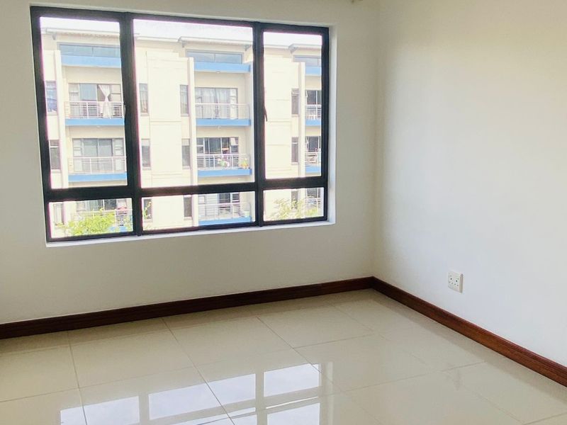 A bright, empty bedroom featuring polished tiled flooring, neutral walls, and a large window providing natural light and a view of the surrounding apartment complex.