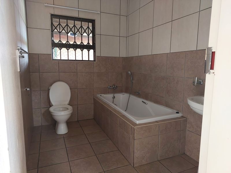 A functional bathroom featuring a built-in bathtub, toilet, and wall-mounted basin with neutral tiled finishes and a security-fitted window.