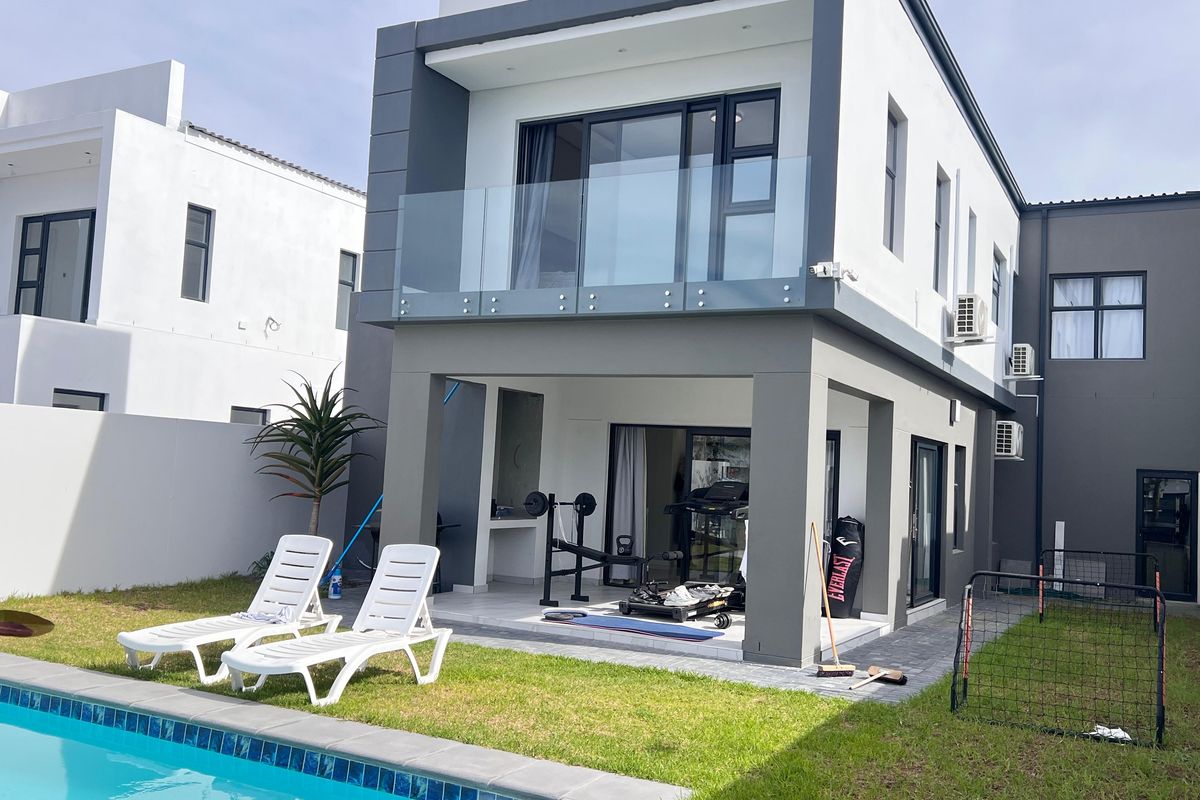 Rear view of a modern double-story home featuring a swimming pool, covered patio with built-in braai, and an upper-level balcony with glass balustrades.