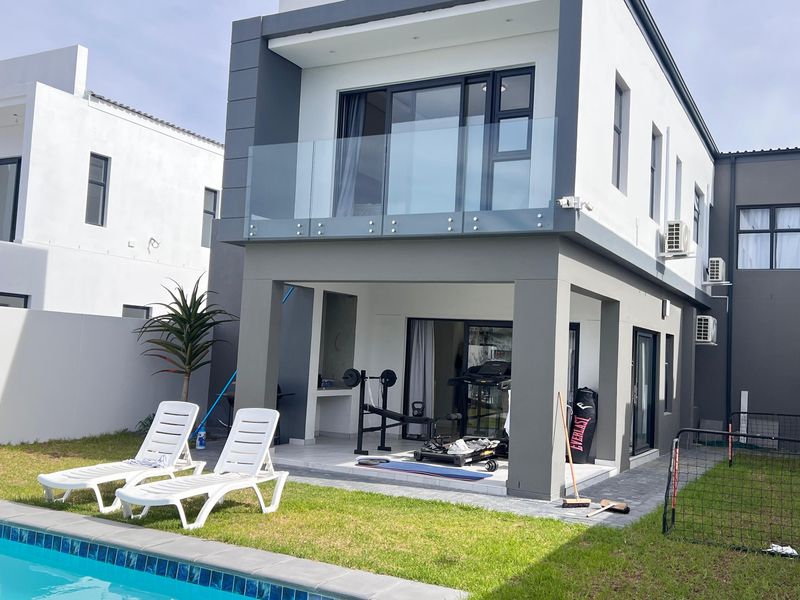 Rear view of a modern double-story home featuring a swimming pool, covered patio with built-in braai, and an upper-level balcony with glass balustrades.