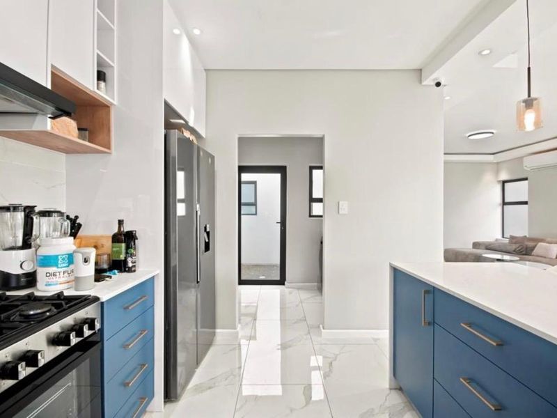 Modern kitchen featuring blue cabinetry, white quartz countertops, marble-look floor tiles, and integrated appliances, leading to a bright open-plan living area.