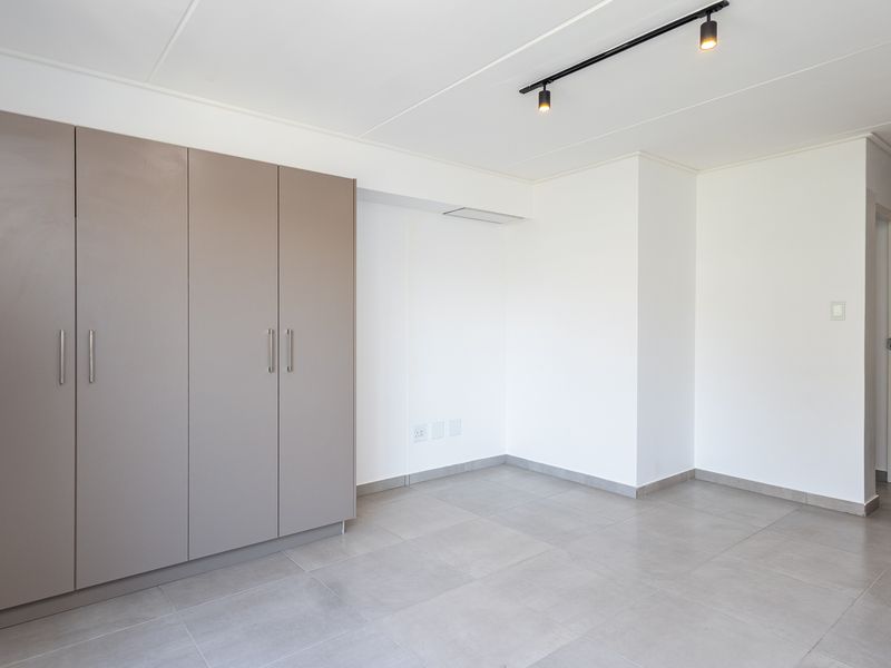 A bright, empty living area featuring neutral tiled flooring, modern track lighting, and ample built-in cupboard storage.