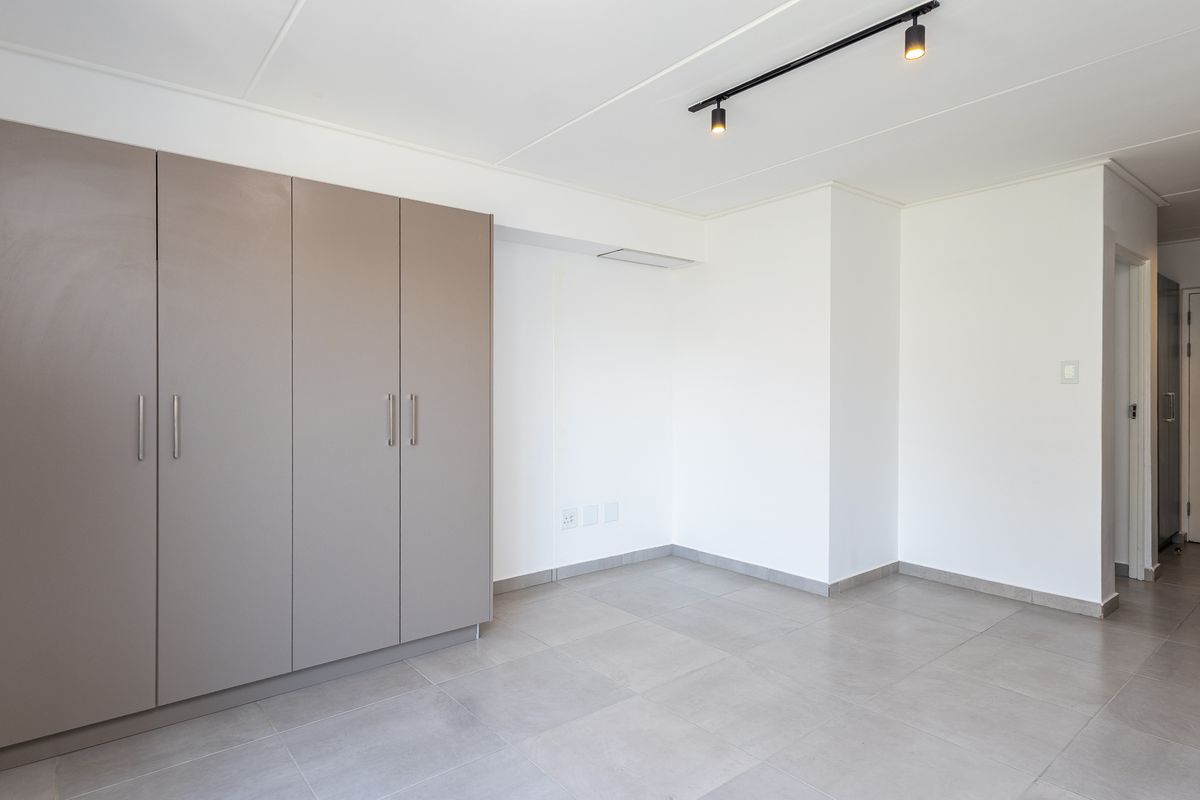 A bright, empty living area featuring neutral tiled flooring, modern track lighting, and ample built-in cupboard storage.
