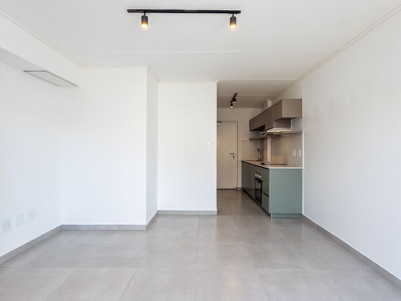 A bright, modern open-plan living and kitchen area featuring neutral tiled floors, contemporary cabinetry, built-in appliances, and minimalist track lighting.