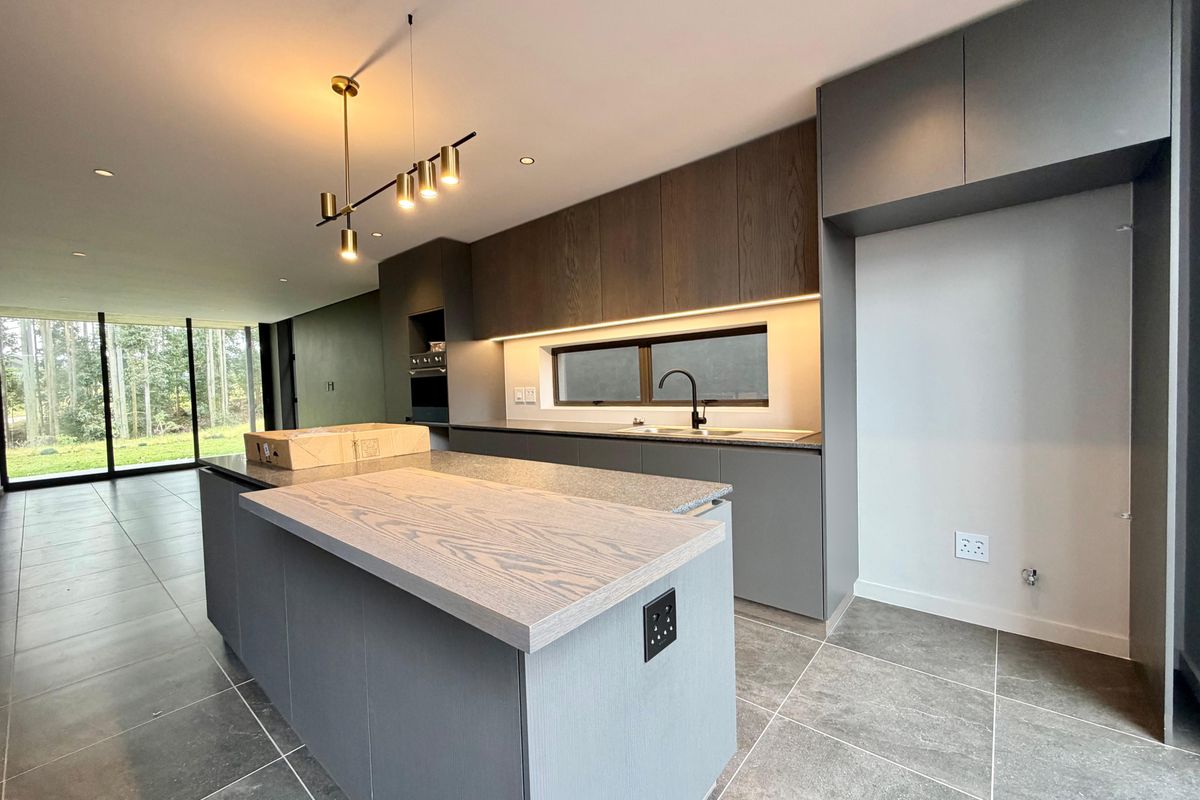Modern kitchen featuring a central island, sleek grey cabinetry, stone countertops, integrated oven, and recessed lighting, with large glass doors leading to the garden.