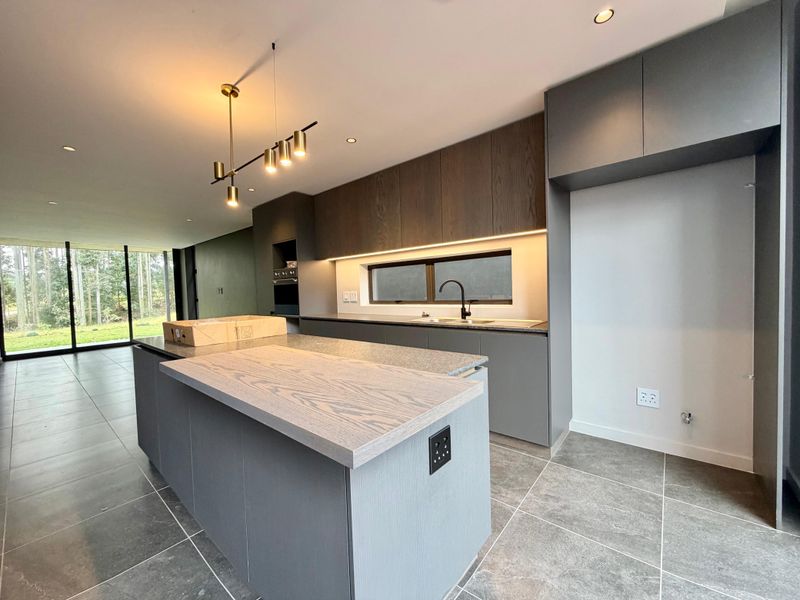 Modern kitchen featuring a central island, sleek grey cabinetry, stone countertops, integrated oven, and recessed lighting, with large glass doors leading to the garden.