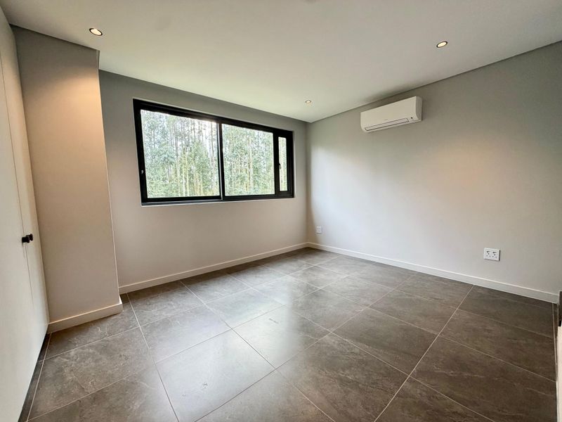A clean, modern, and empty bedroom featuring neutral tiled flooring, recessed lighting, a wall-mounted air conditioning unit, and a built-in cupboard.