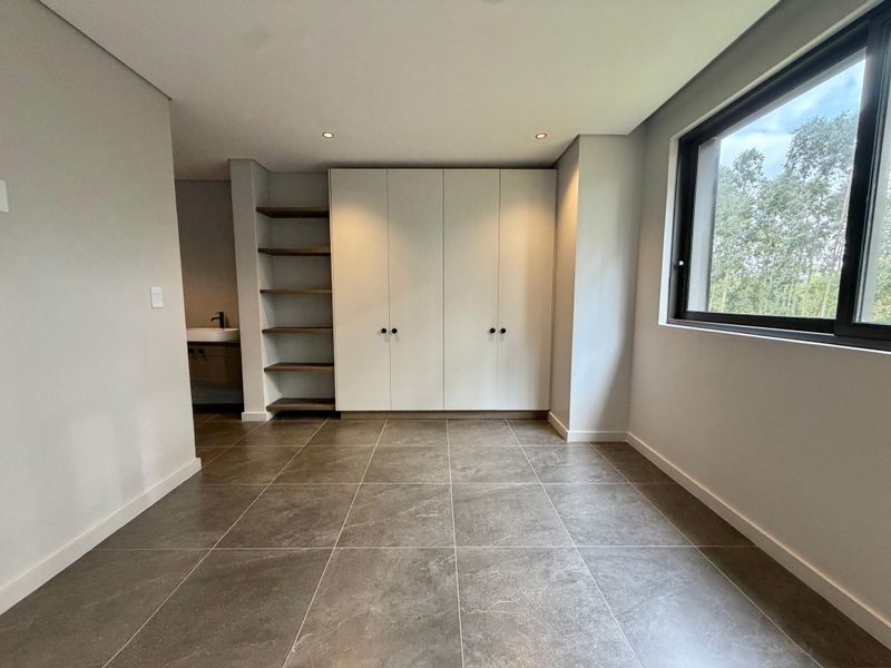A modern, empty bedroom featuring tiled floors, built-in cupboards, open shelving, and an integrated vanity area with a view of the surrounding trees.