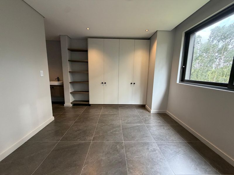 Modern bedroom featuring large format floor tiles, ample built-in cupboard space, open shelving, and an integrated vanity area with a window overlooking greenery.
