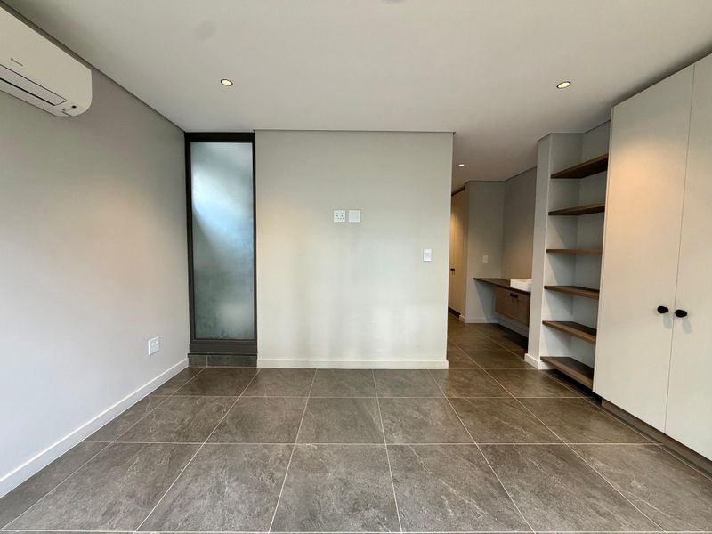 Modern, spacious bedroom featuring large format floor tiles, built-in storage, integrated shelving, and an en-suite vanity area with a split-system air conditioner.