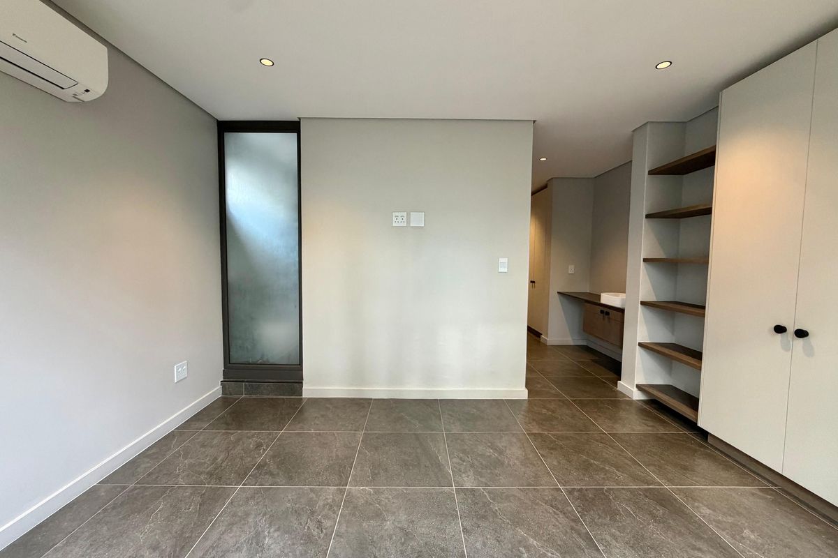 Modern, spacious bedroom featuring large format floor tiles, built-in storage, integrated shelving, and an en-suite vanity area with a split-system air conditioner.