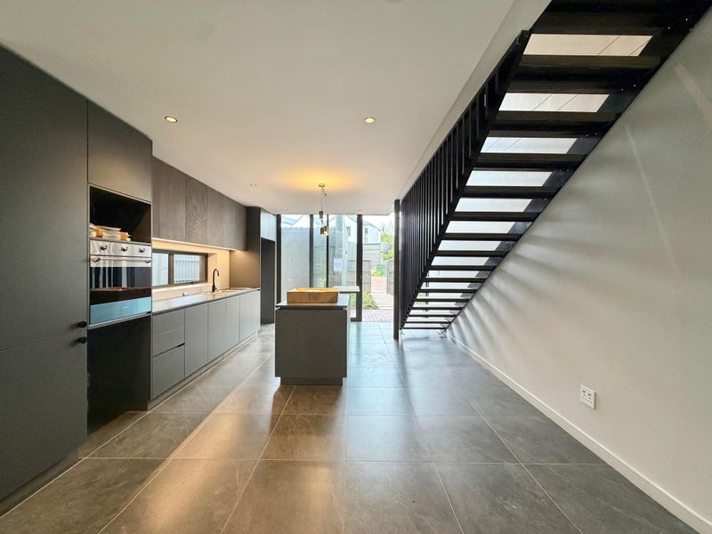 Modern open-plan kitchen featuring sleek dark cabinetry, a central island, and a striking architectural staircase, leading to a rear outdoor area.