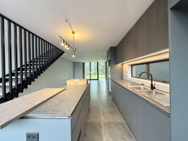 Modern kitchen featuring sleek dark cabinetry, stone countertops, integrated lighting, and a striking staircase, leading to an open-plan living area with garden views.