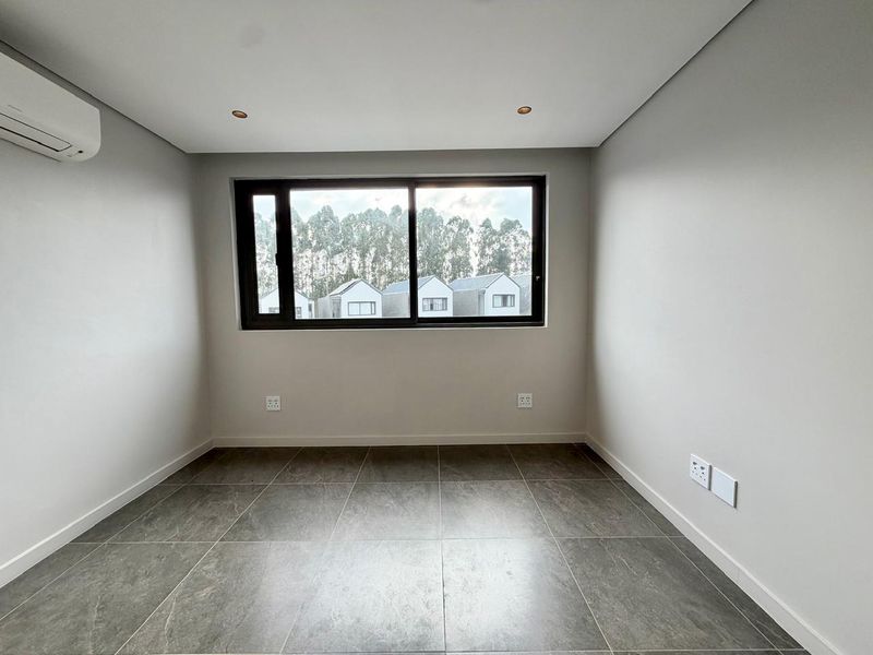A clean, modern, and empty bedroom featuring neutral tiled flooring, recessed lighting, and a wall-mounted air conditioning unit with a view of the neighborhood.