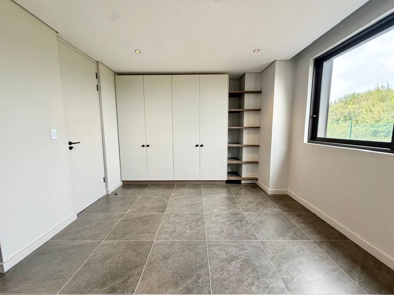 A bright, modern bedroom featuring neutral tiled flooring, ample built-in cupboard space, integrated open shelving, and large windows providing natural light.