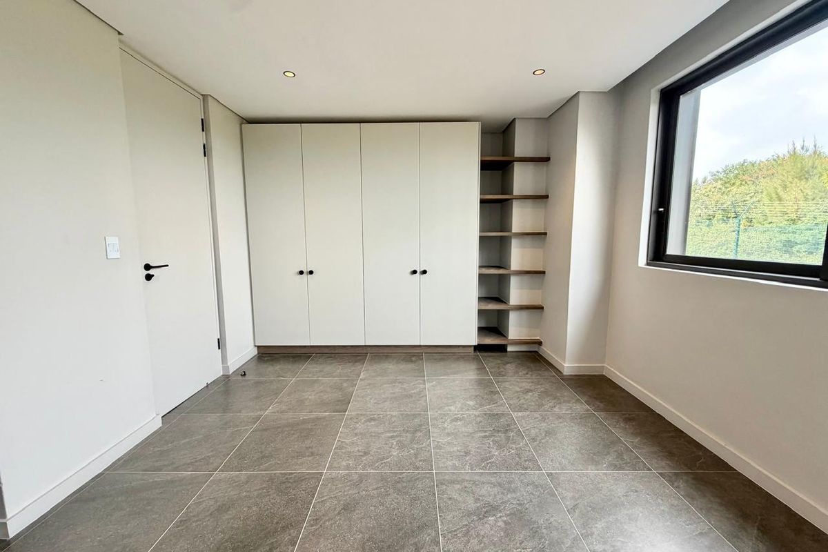 A bright, modern bedroom featuring neutral tiled flooring, ample built-in cupboard space, integrated open shelving, and large windows providing natural light.