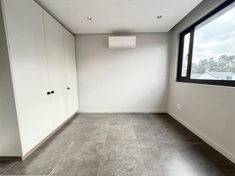 A clean, modern bedroom featuring neutral tiled flooring, built-in storage cupboards, and a wall-mounted air conditioning unit.