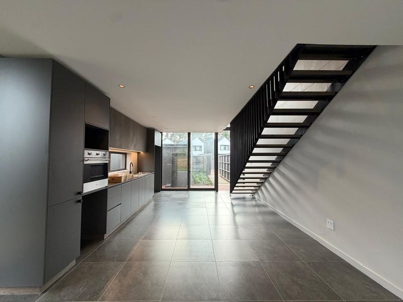 Modern open-plan kitchen featuring sleek dark cabinetry, integrated appliances, and large format floor tiles, leading to a bright outdoor area via sliding glass doors.