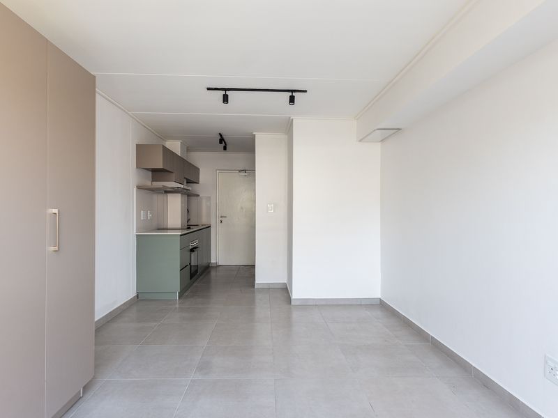 A bright, open-plan living area featuring modern tiled flooring, track lighting, and a compact kitchen with built-in cabinetry and integrated appliances.