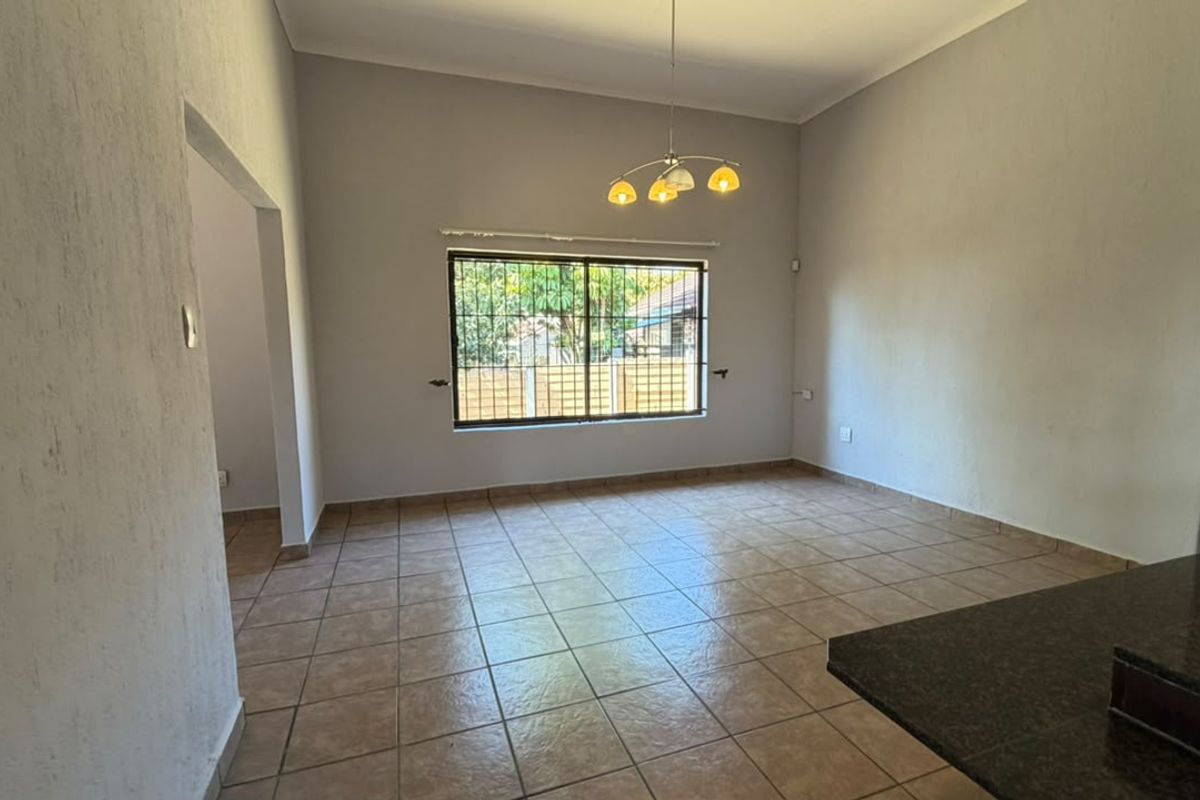 An empty, open-plan living area featuring neutral tiled flooring, a large window with security bars, and a modern pendant light fixture.